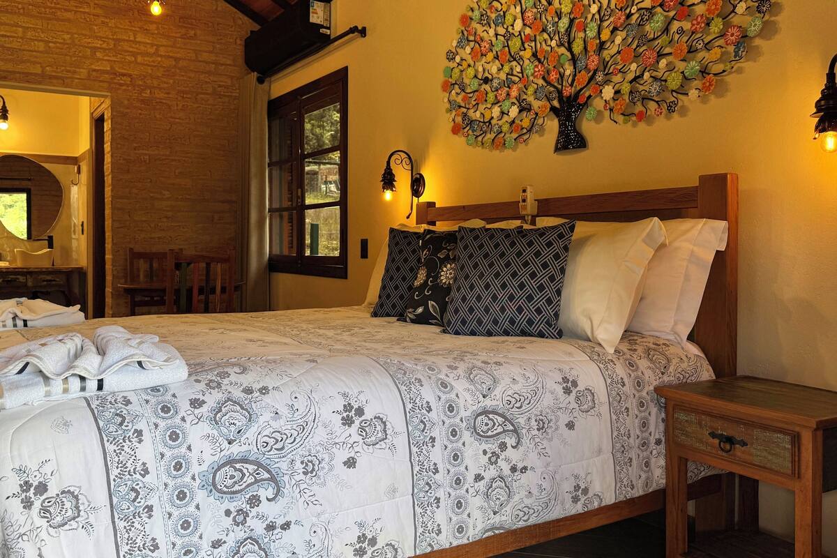 A neatly made bed with patterned bedding, a wooden nightstand, a wall-mounted flower art piece, and a mirror above a wooden dresser.
