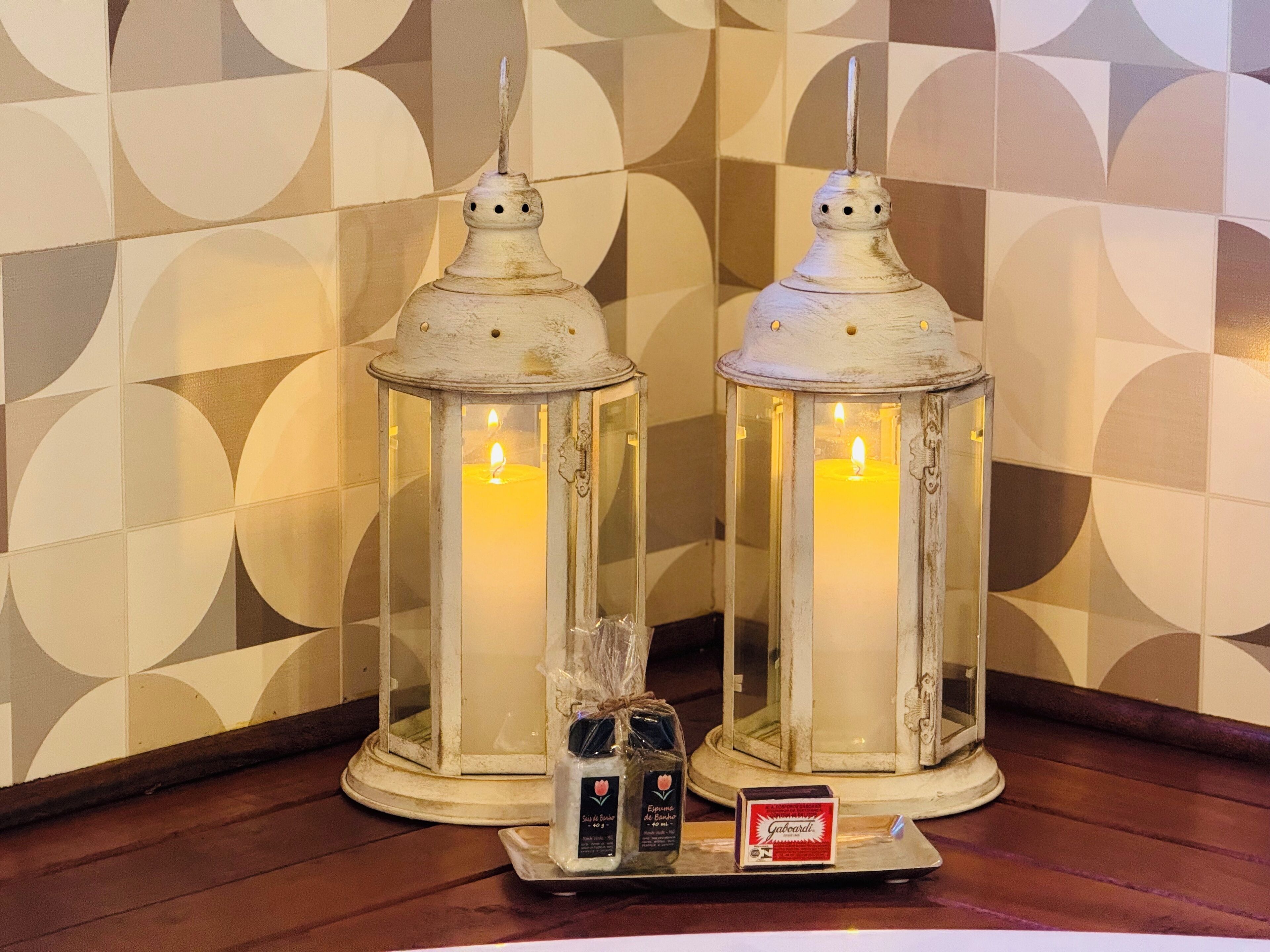 Dos faroles blancos de estilo vintage con velas, sobre una bandeja con botellas pequeñas y una caja, frente a una pared con un diseño geométrico.