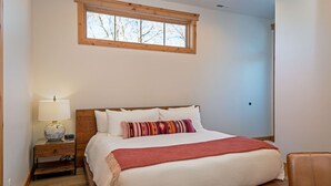 A bedroom with a wooden bed, white bedding, a red throw blanket, a nightstand with a lamp, and a window with a view of bare trees. - New! Condo Downtown with rooftop patio, steam shower, mountain views (Whitefish)