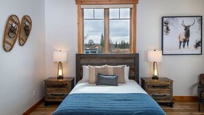 Een slaapkamer met een groot bed, een houten hoofdeinde, twee nachtkastjes met lampen, een raam met uitzicht op huizen, en twee decoratieve objecten aan de muur.
