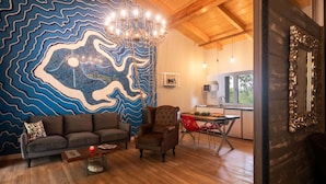 A room with a blue wavy wall mural, a wooden ceiling, a sofa, a chair, a dining table with red chairs, and a kitchen area visible through an open door.