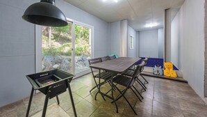 Un espace repas comprenant une grande table, des chaises, un coin barbecue et une vue sur un espace extérieur aménagé, visible à travers des portes vitrées.