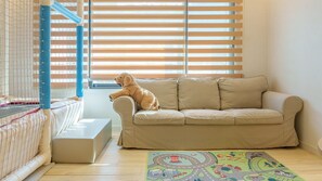 Ein Zimmer mit einem beigen Sofa, einem Reisebettchen und einem farbenfrohen Teppich.