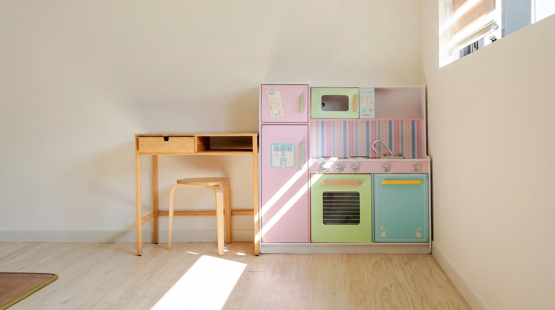 A room with a wooden desk, a stool, and a colorful play kitchen.