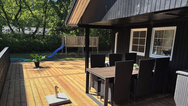 Een houten terras met een tafel, stoelen, een kleine plant en een glijbaan op de achtergrond.