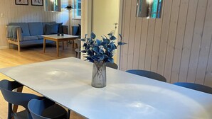 Een eethoek met een lange tafel, vier stoelen, een vaas met bloemen, hanglampen en een houten vloer.