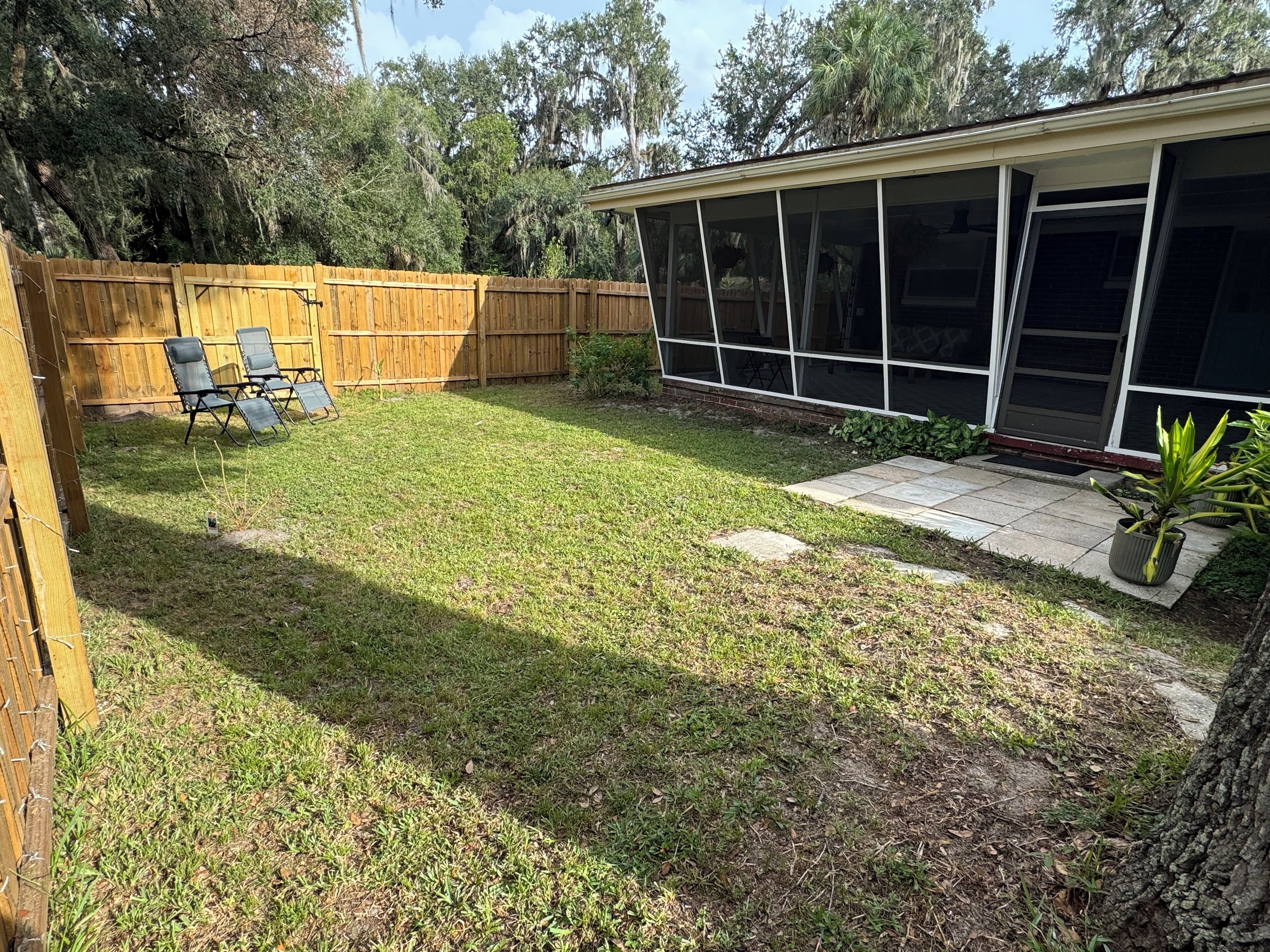 A backyard with a wooden fence, a patio with chairs, and a house with large windows.