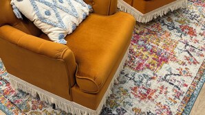 Two mustard-colored armchairs with tufted backs and decorative pillows, placed on a colorful patterned rug in a room with wooden flooring and a teal door.