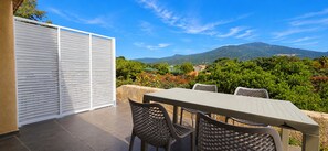 Une terrasse extérieure avec une table et des chaises, une porte blanche à persiennes et une vue sur une montagne.