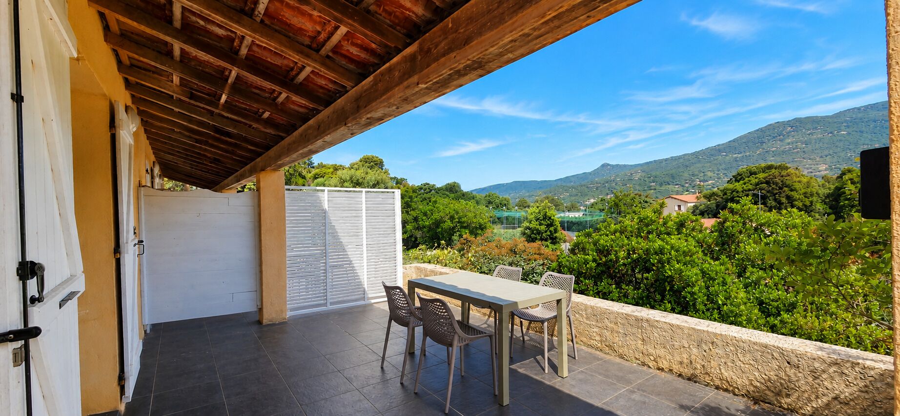 Une terrasse couverte avec une table et des chaises, une vue sur un paysage montagneux et une clôture blanche.