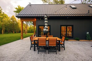 A patio with a table and chairs, a hanging umbrella, and a building with large windows and a metal roof. - Stillwater Retreat | Pickleball Court + Private Pond w/ Kayaking (Big Prairie)
