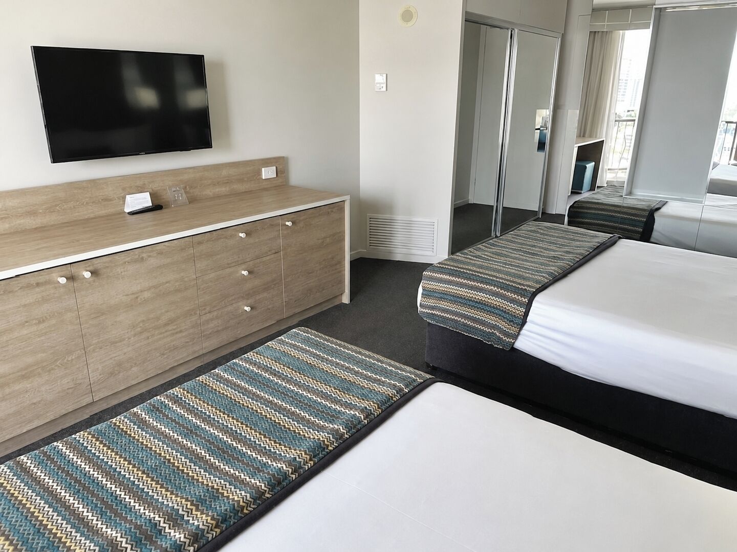 Ein Hotelzimmer mit Flachbildfernseher, einem Holzkoffer, einem Bett mit gestreifter Bettdecke, einer Bank und einem Spiegel.