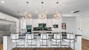 Cocina moderna con muebles blancos, una isla central con taburetes de bar, electrodomésticos de acero inoxidable y una araña.