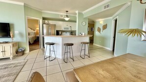 A modern kitchen with white cabinets, a central island with stools, a dining area with a wooden table, and a living room with a TV.