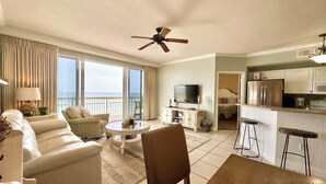A spacious living room with a sofa, armchair, coffee table, and a TV. There's a balcony with a view of the ocean.