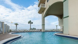 A swimming pool with a curved edge, surrounded by a low wall and a covered area with a balcony.