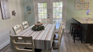 Un coin repas avec une nappe à rayures, des chaises en bois et une vue sur l’extérieur à travers des portes vitrées.