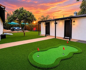 A golf course with a putting green, a patio area with seating, and a building with doors and windows.