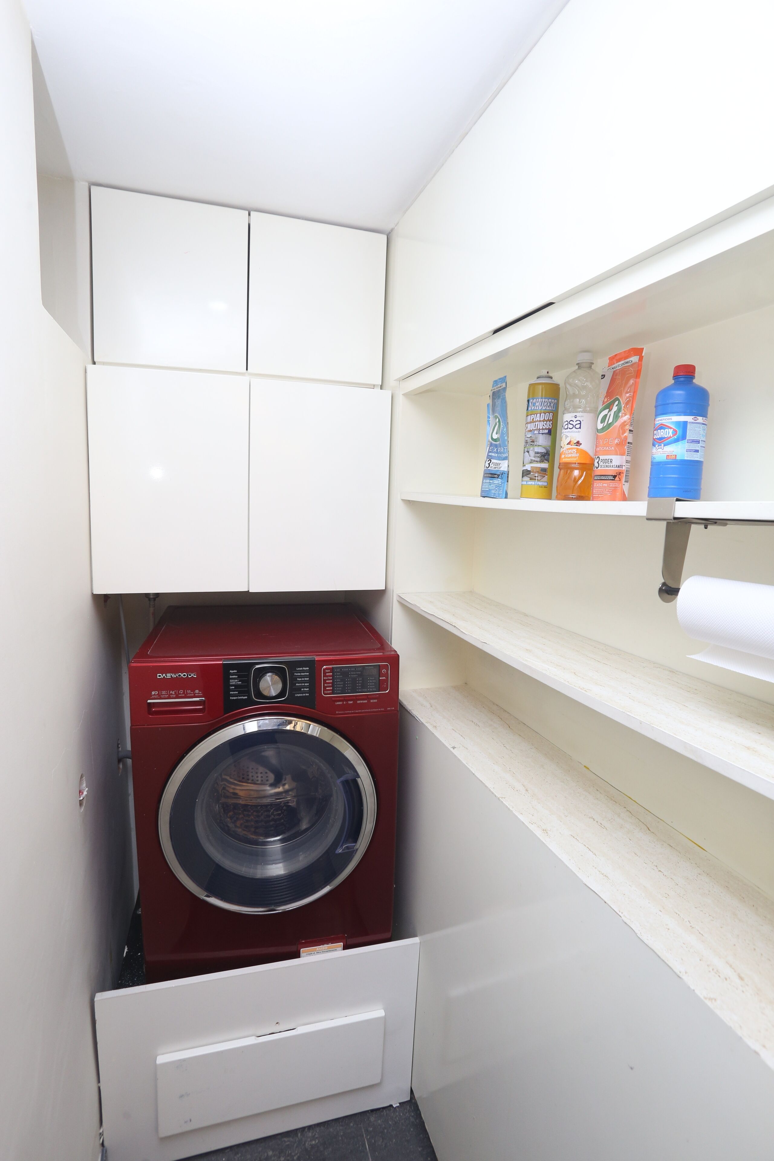 Une petite buanderie avec un lave-linge rouge, des placards blancs et des étagères où sont rangés les produits d’entretien.