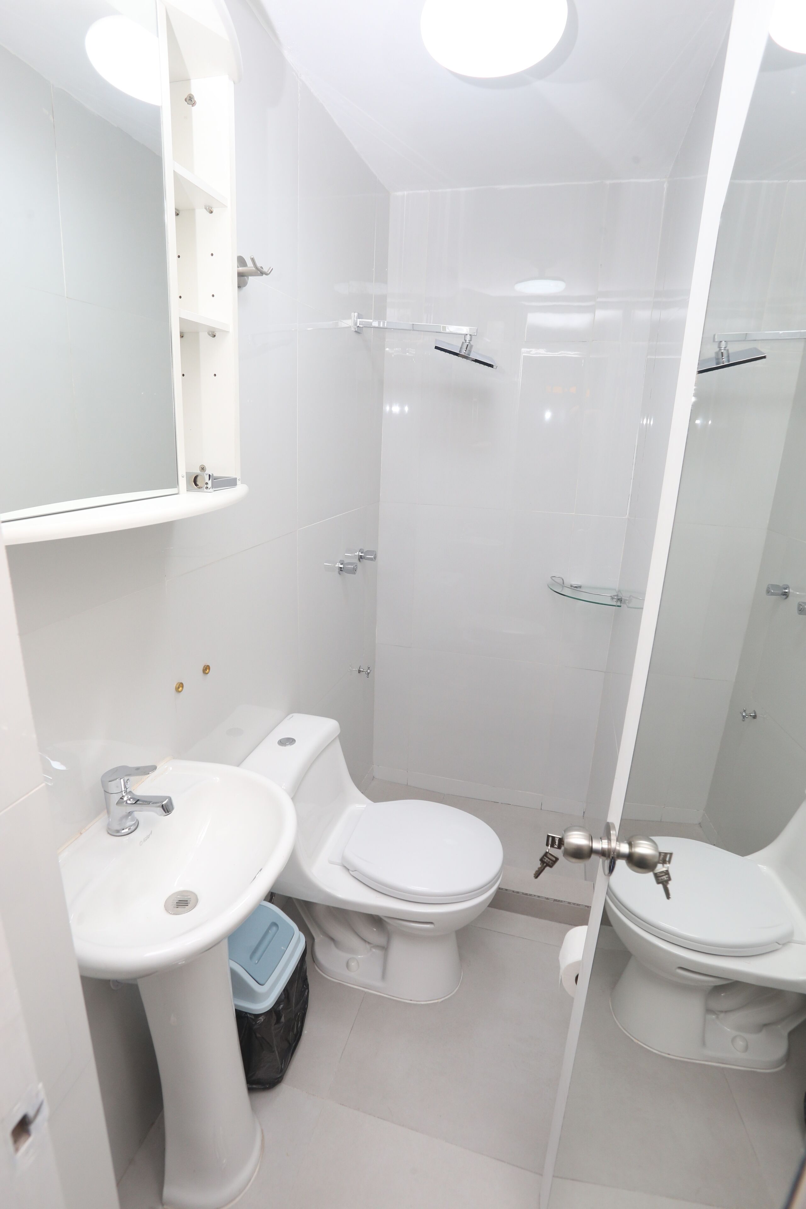 Un bany modern amb un lavabo de pedestal, dues vàteres, un espai de banyera amb portes de vidre i un mirall sobre el lavabo.
