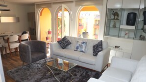 Una sala de estar moderna con un sillón gris, un sofá blanco con cojines estampados, una mesa de centro de vidrio y un otomano gris.