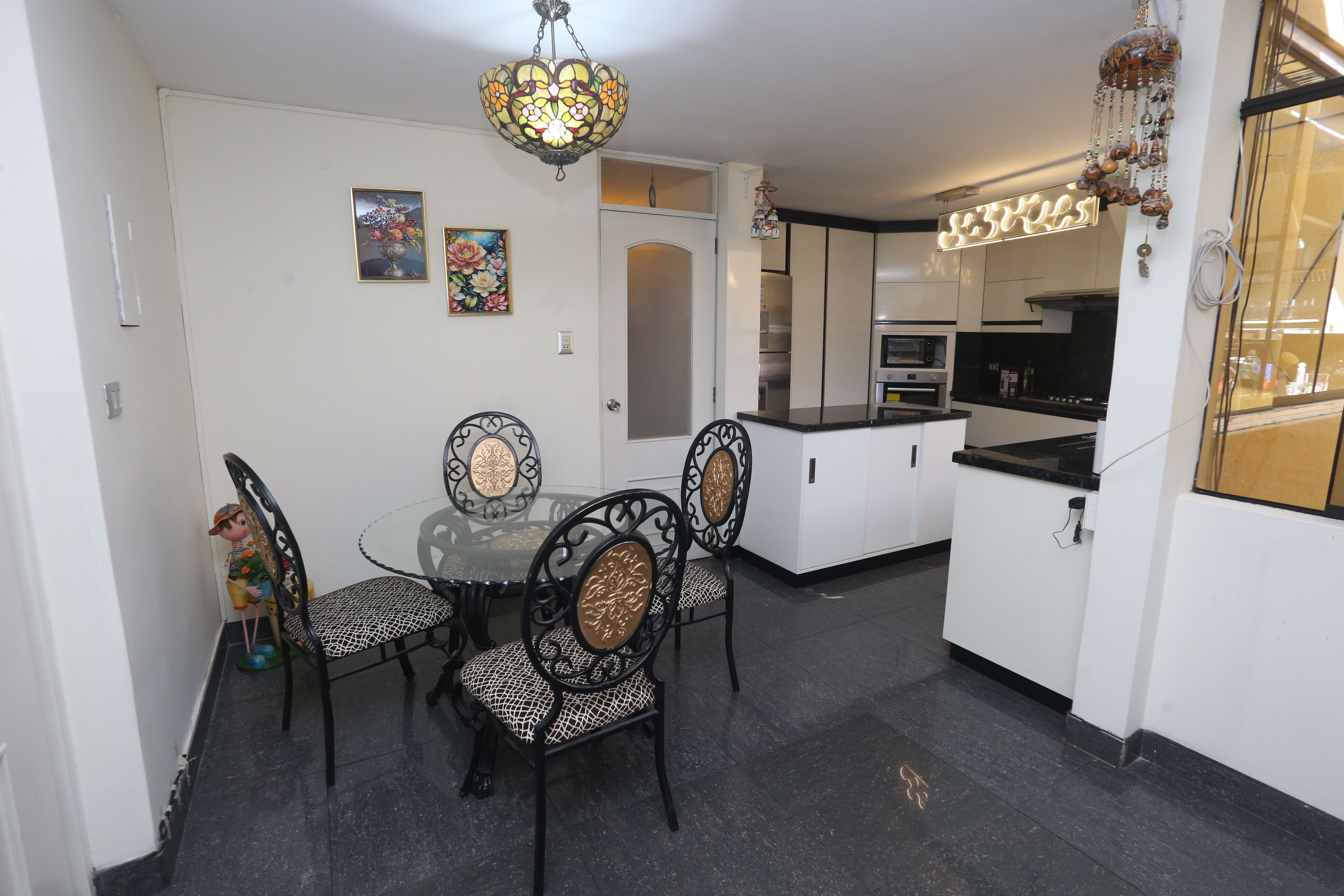 A dining area with a glass table and black chairs with gold accents, a chandelier, and a kitchen in the background.