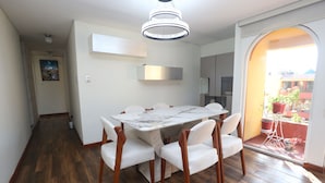 Un área de comedor con una mesa de mármol y sillas blancas, una lámpara moderna y vistas a un balcón exterior con plantas en macetas a través de un arco.