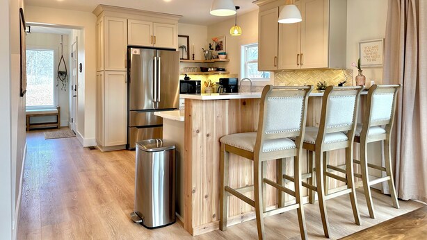 A modern kitchen with stainless steel appliances, wooden bar stools, and a trash bin.