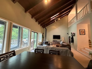 A spacious living area with a large wooden beam ceiling, a fireplace, a flat-screen TV, and a staircase leading to an upper level.
