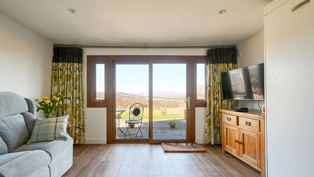 Een woonkamer met een bank, een televisie op een houten kast en een schuifdeur naar een terras met een stoel en een tafel.