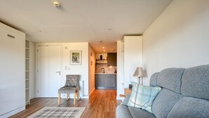 Een moderne woonkamer met een grijze bank, een geruit vloerkleed en een fauteuil. Achtergrond toont een keuken.