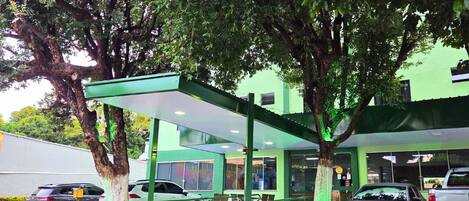 A covered outdoor area with a car parked under a tree, a building with a green facade in the background, and a paved ground.
