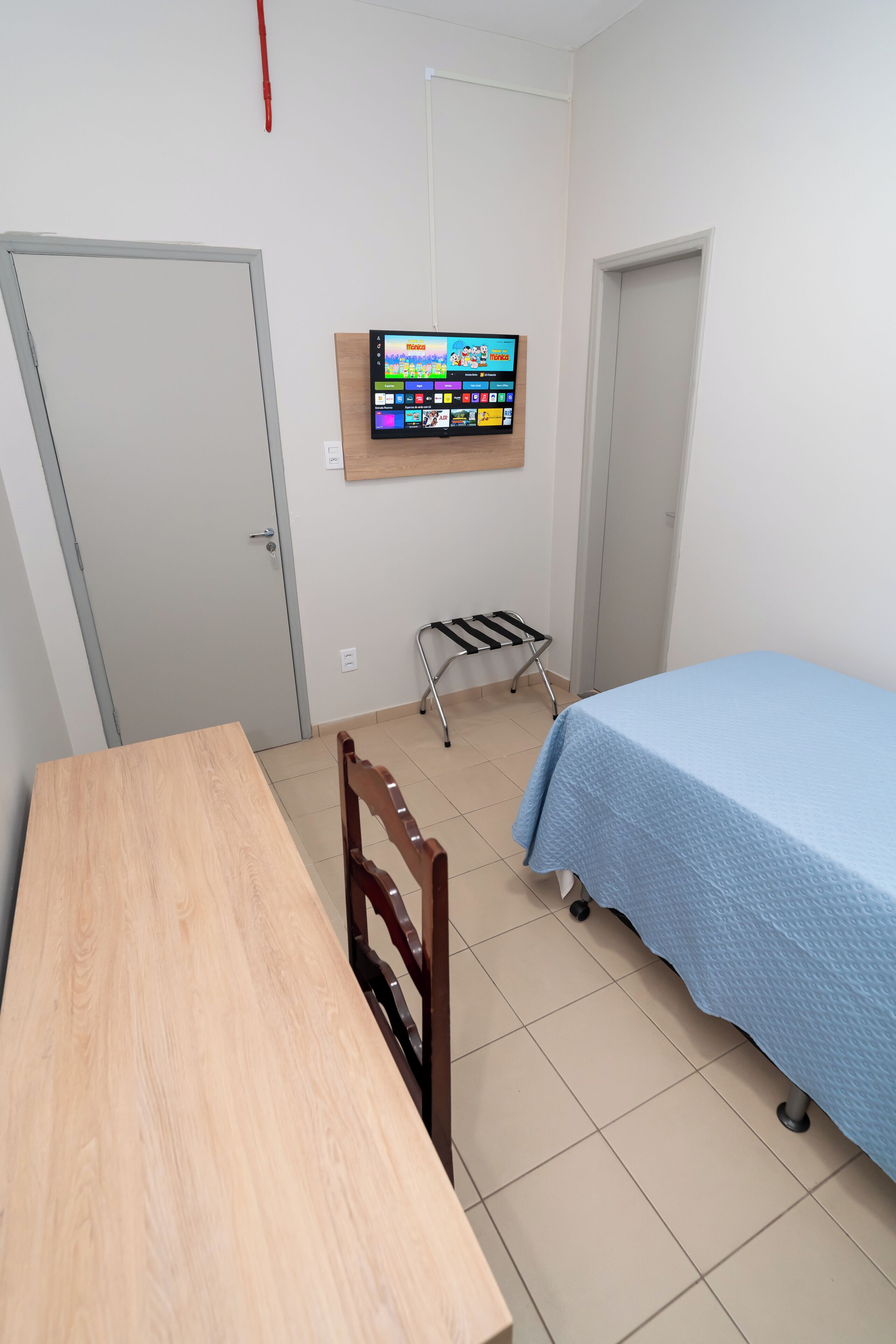 Una stanza con un tavolo in legno, un letto blu, una TV a muro e una piccola sedia.