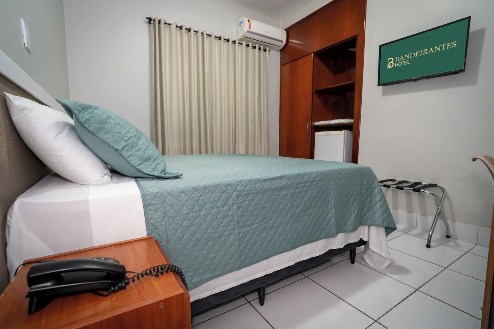 Una habitación de hotel con una cama, un teléfono en la mesita de noche, un radiador y un cartel con el nombre del hotel.