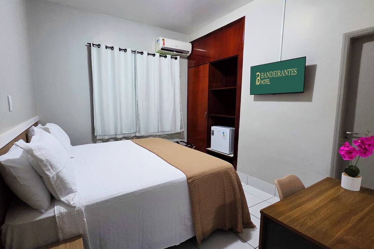 A hotel room with a bed, a desk, a chair, a wardrobe, an air conditioning unit, a refrigerator, and a sign indicating the hotel name.