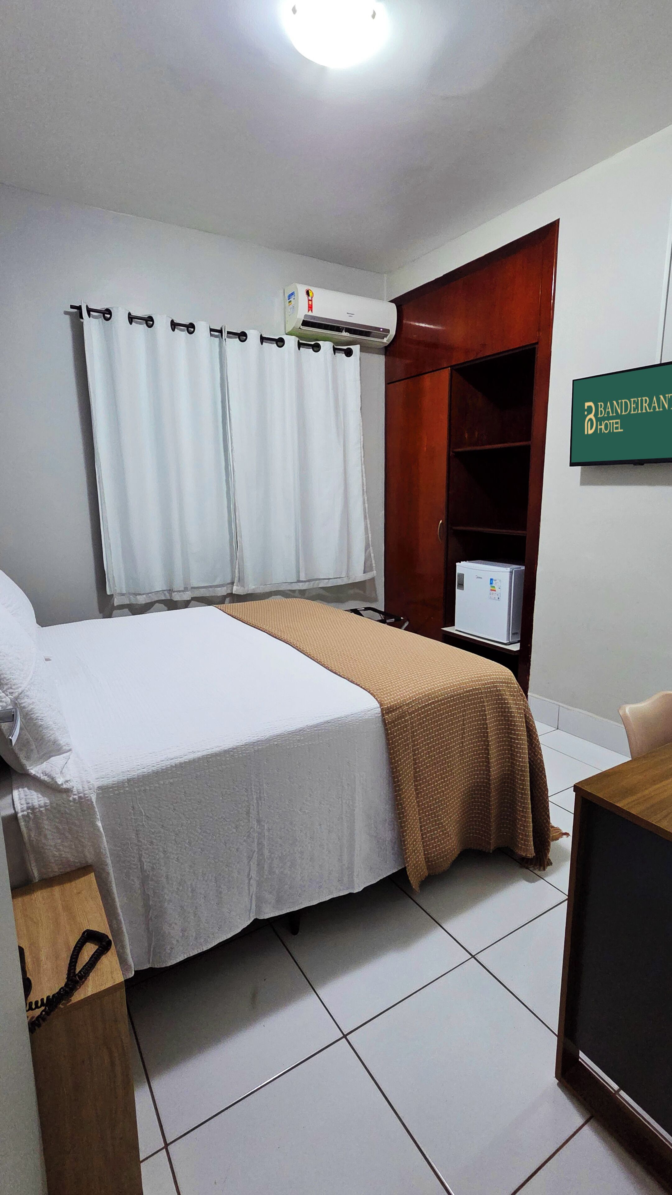 A hotel room with a bed, an air conditioner, a wooden wardrobe, and a desk with a computer.