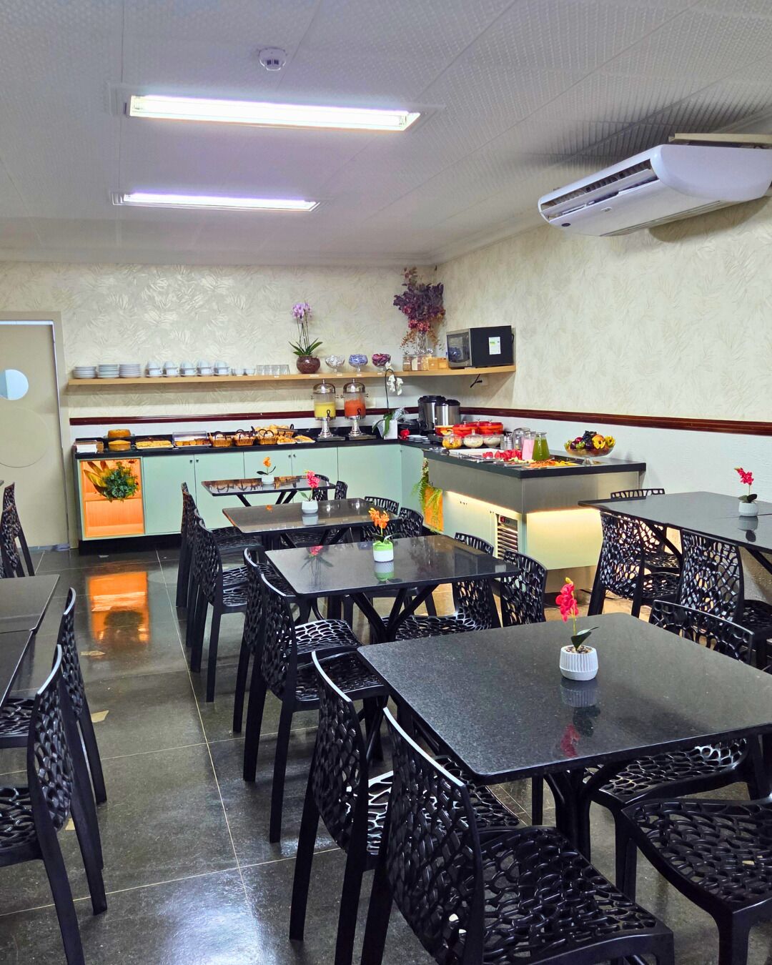 A restaurant interior with black metal chairs and tables, a kitchen counter with various food items, and an air conditioning unit on the ceiling.