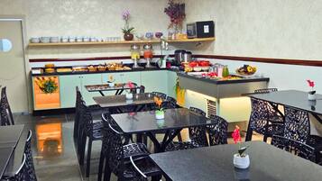 A restaurant interior with black metal chairs and tables, a kitchen counter with various food items, and an air conditioning unit on the ceiling.