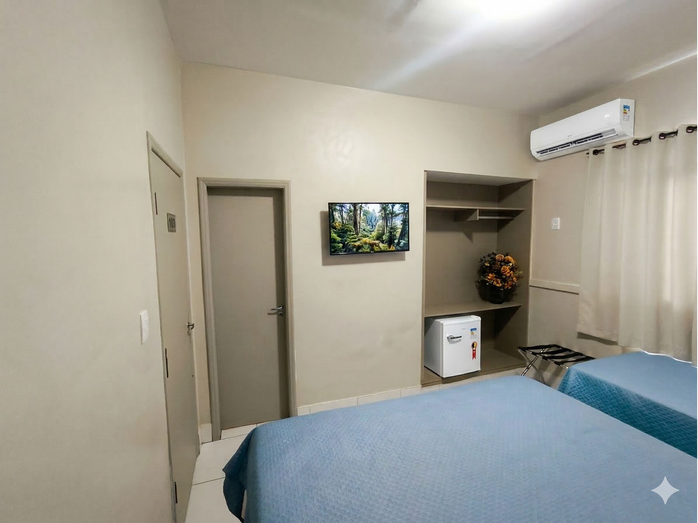 A hotel room with a bed, a TV, a small fridge, and an air conditioning unit.