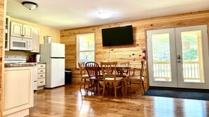 Una cocina compacta con armarios de madera, un microondas y un refrigerador. Hay una mesa de comedor con sillas y un televisor de pantalla plana montado en la pared.