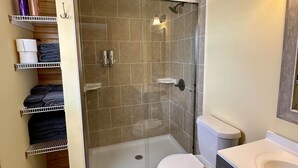 Un baño con ducha de vidrio, inodoro, lavamanos y repisas con artículos de aseo.