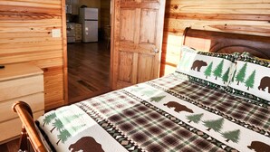 Interior de una cabaña de madera con una cama, un aparador de madera y una cocina visible a través de una puerta abierta.