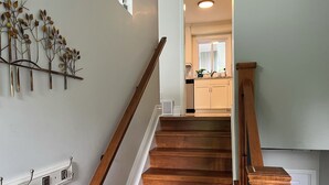 A wooden staircase with a handrail, a wall-mounted decorative metal piece, and a kitchen area in the background. - Cozy Countryside Retreat (Clarence-Rockland)