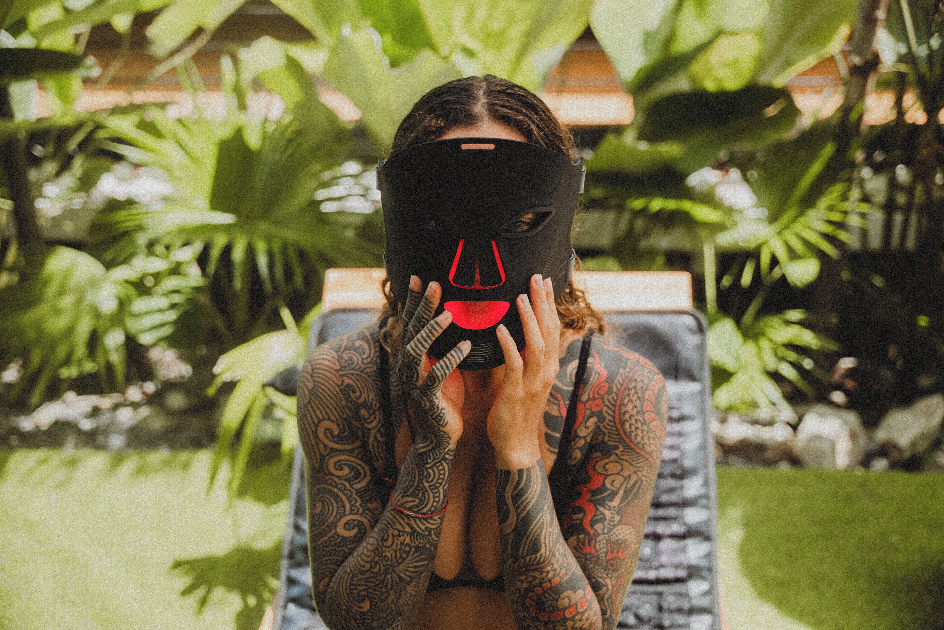 A person with tattoos sitting in a chair outdoors with greenery in the background.