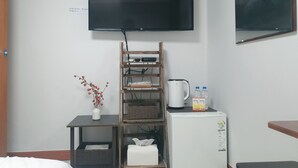 A hotel room with a flat-screen TV mounted on the wall, a wooden shelf with baskets, a small refrigerator, and a bedside table with a vase of flowers. - Liebe stay (Busan)