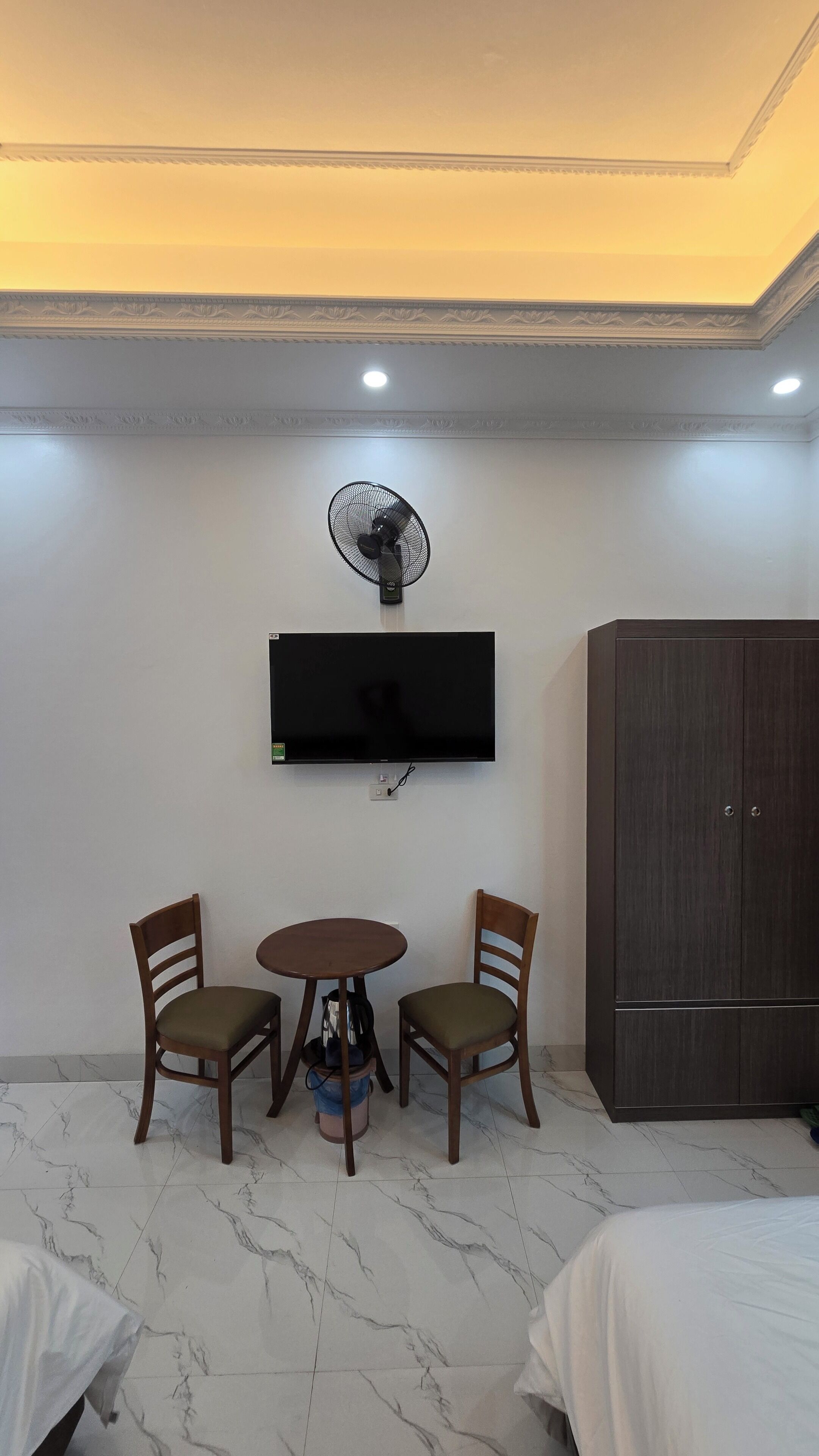 A hotel room with a flat-screen TV mounted on the wall, a ceiling fan, a wooden wardrobe, two wooden chairs, a small round table, and a marble-tiled floor.