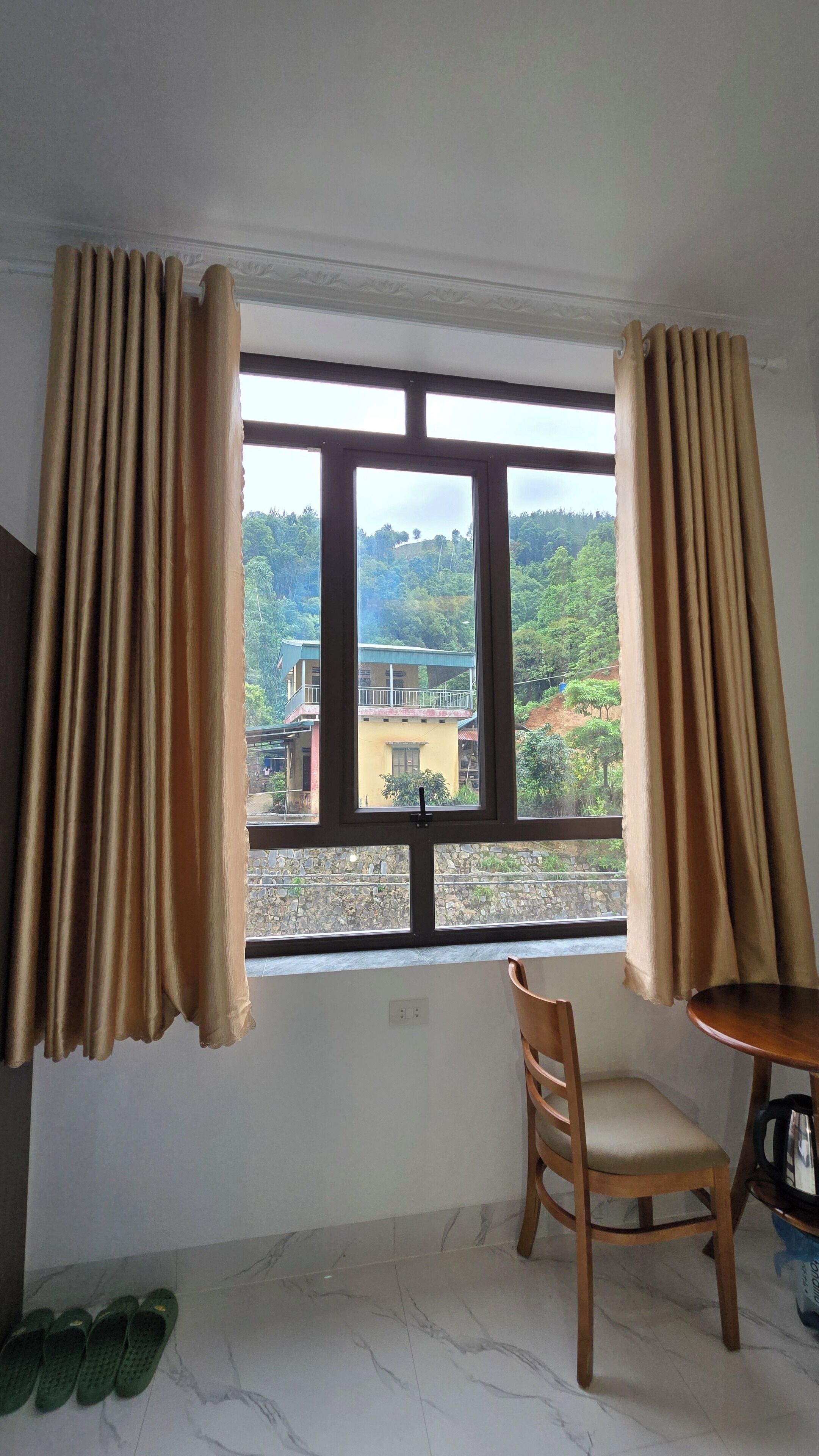 Ein Zimmer mit einem großen Fenster, einem Holzstuhl, einem kleinen Tisch und einem Paar Hausschuhen auf dem Boden.