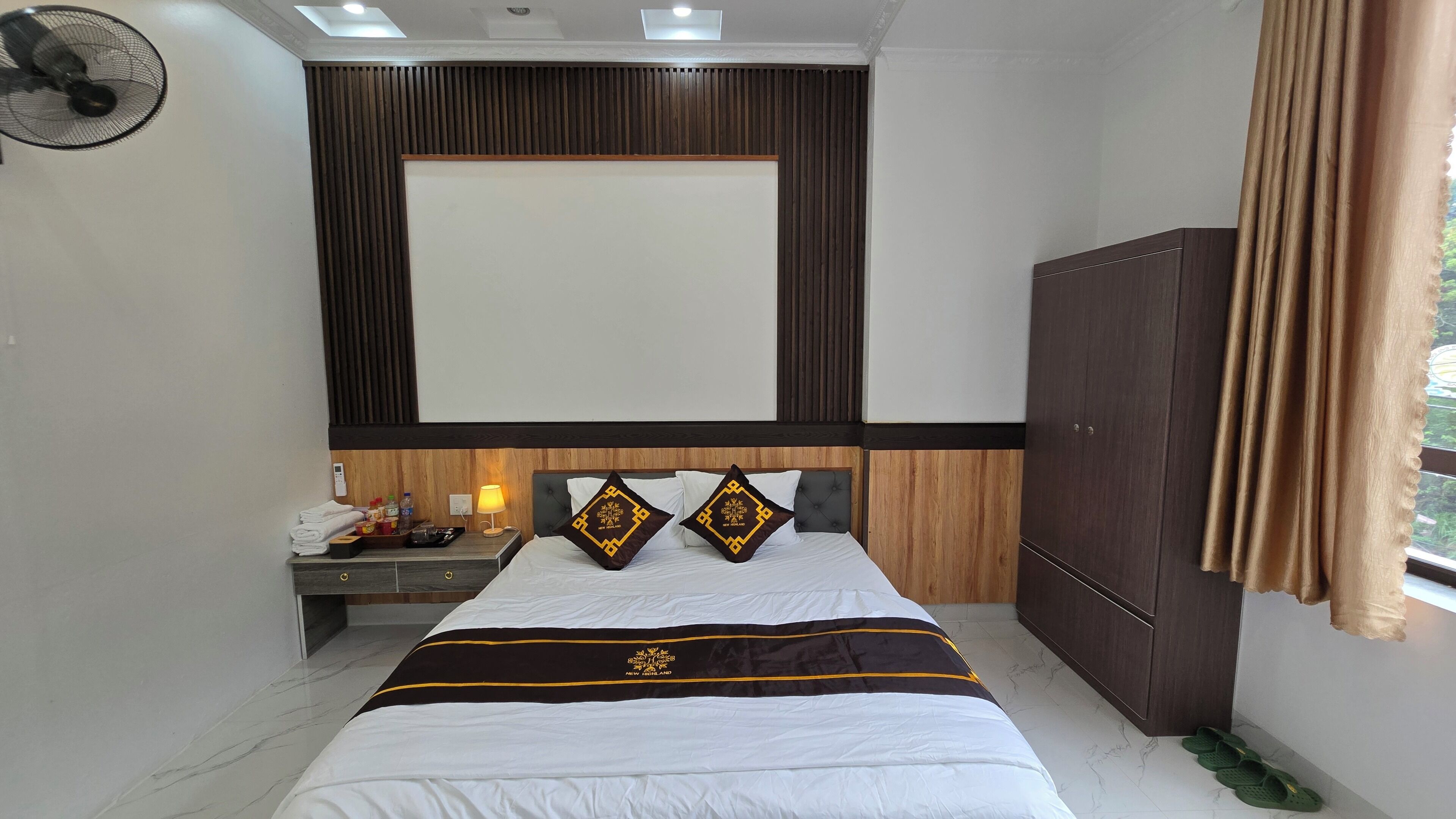 A hotel room with a bed, a nightstand, a lamp, a fan, and a wardrobe.