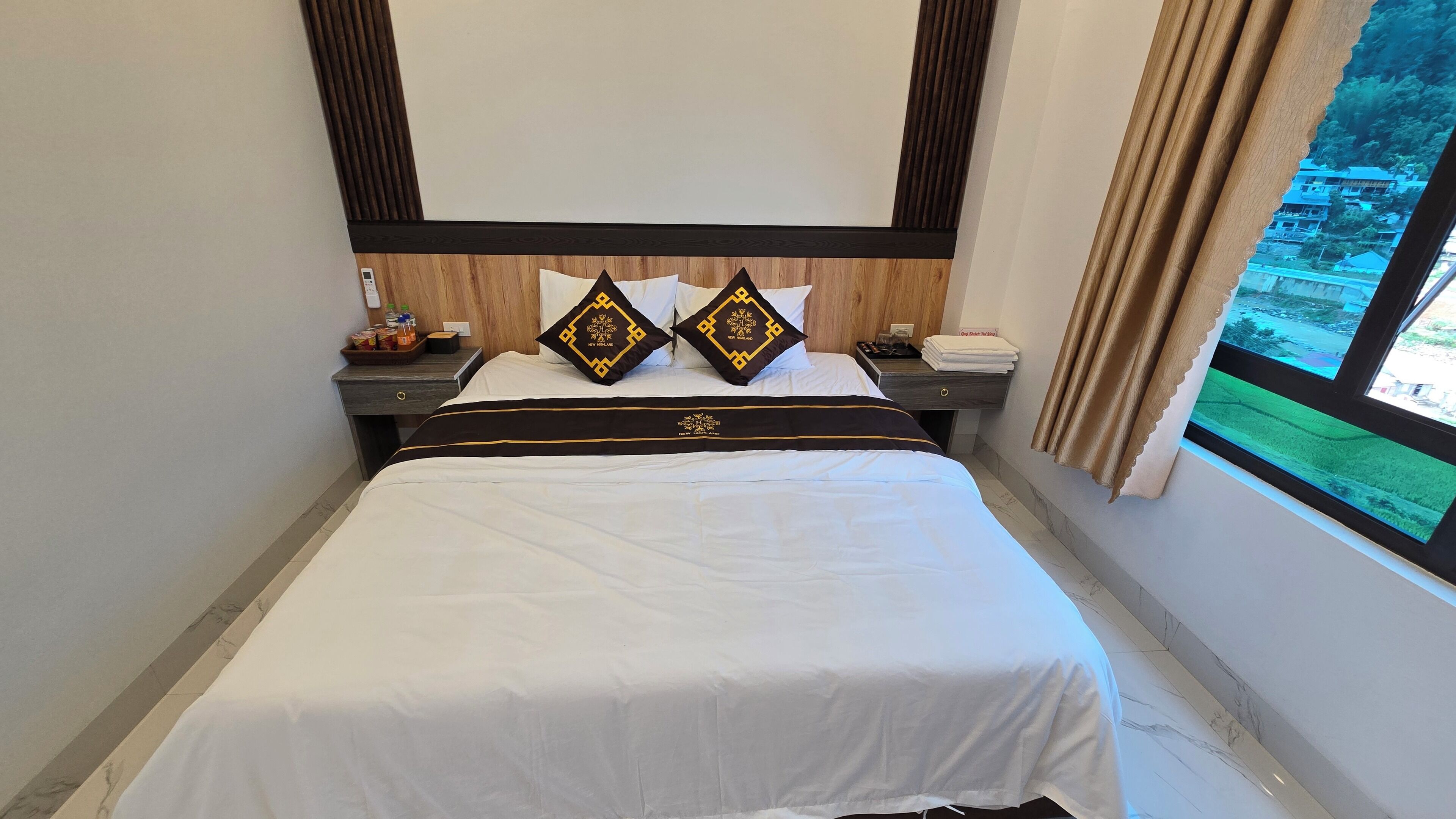 Een hotelkamer met een bed, twee nachtkastjes, een houten hoofdeinde en een raam met uitzicht op groen.