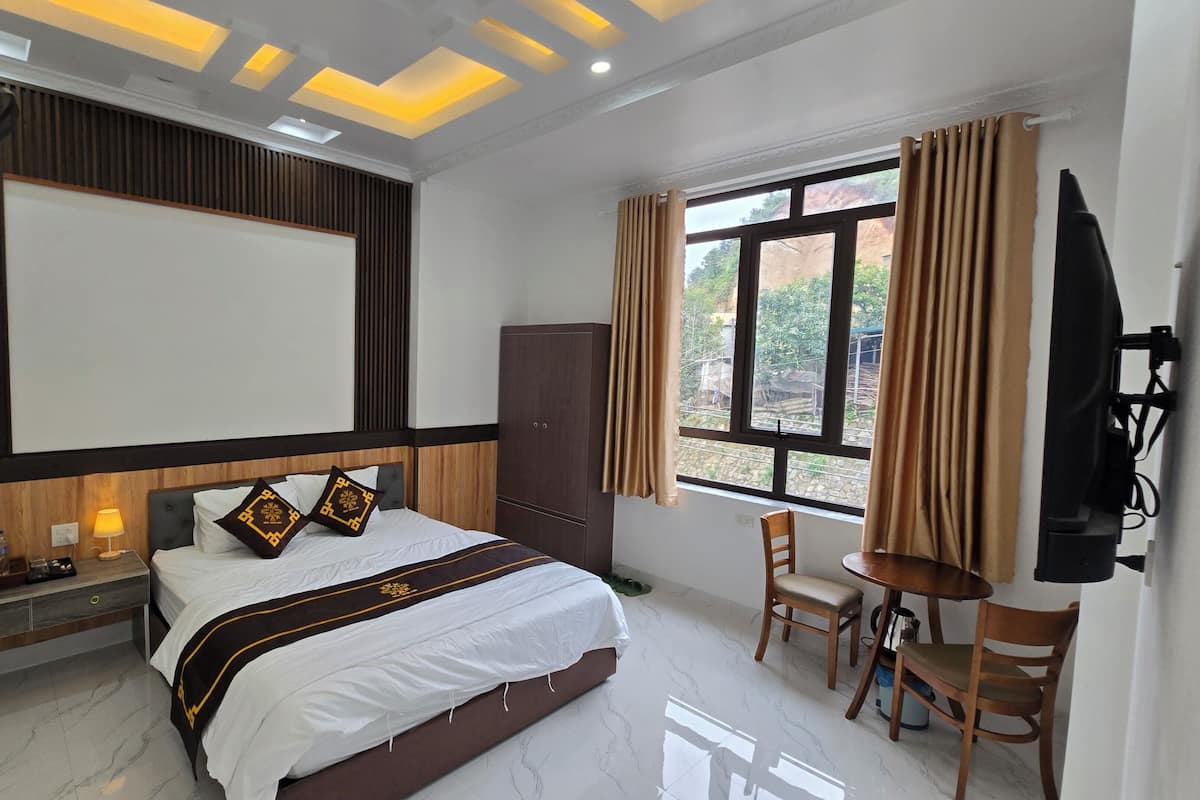 A hotel room with a bed, bedside table, lamp, wardrobe, TV, and a window with curtains.